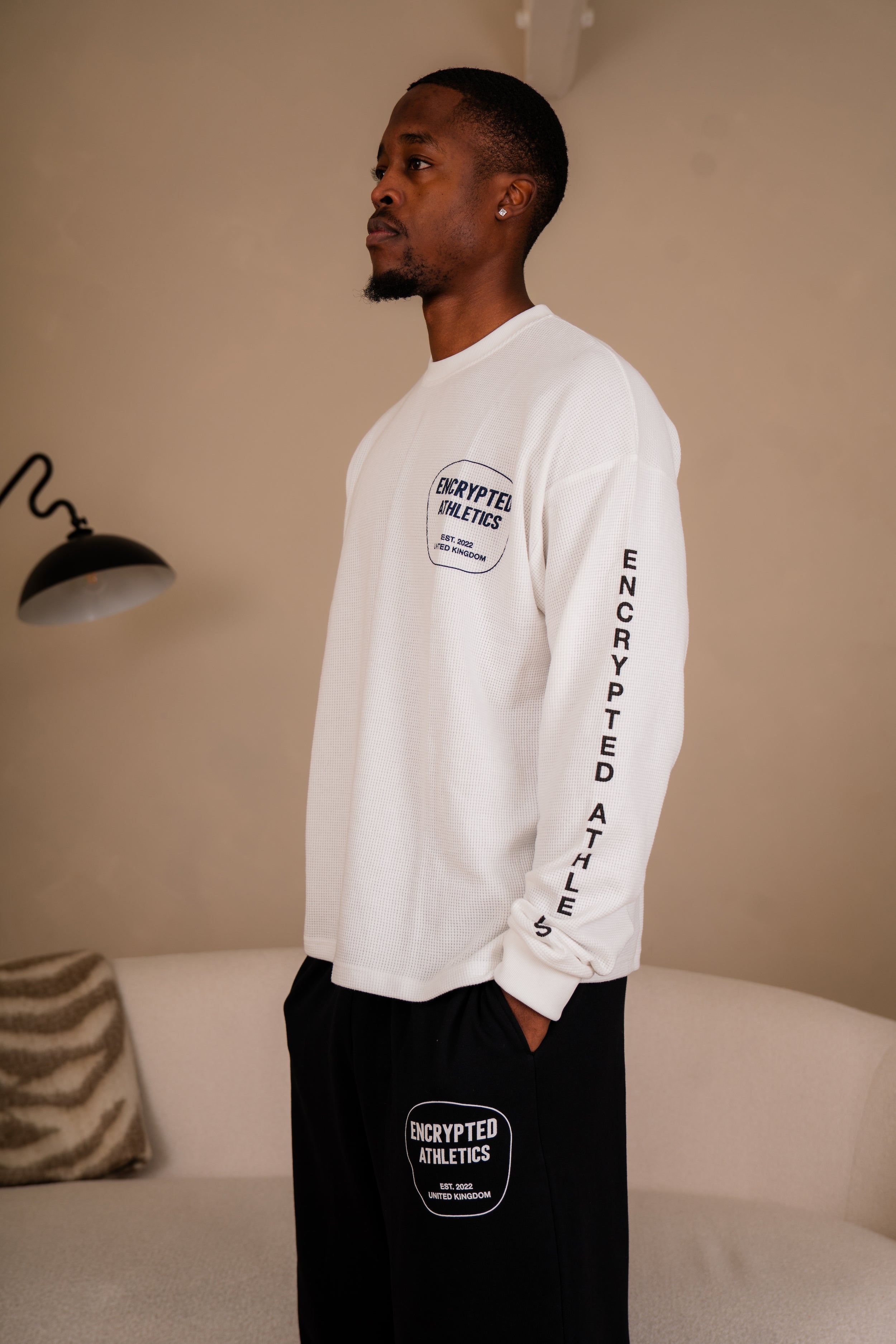 Man wearing a white long-sleeve shirt and black pants with 'Encrypted Athletics' branding in a room.