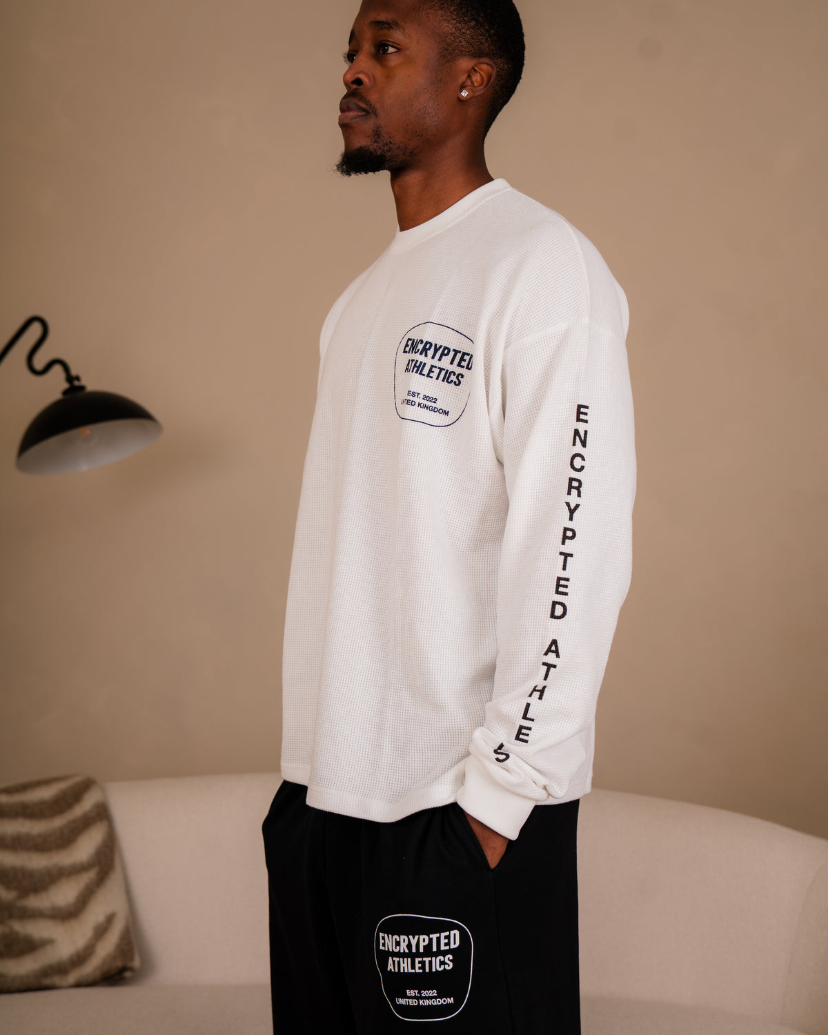 Man wearing a white long-sleeve shirt and black pants with 'Encrypted Athletics' branding in a room.